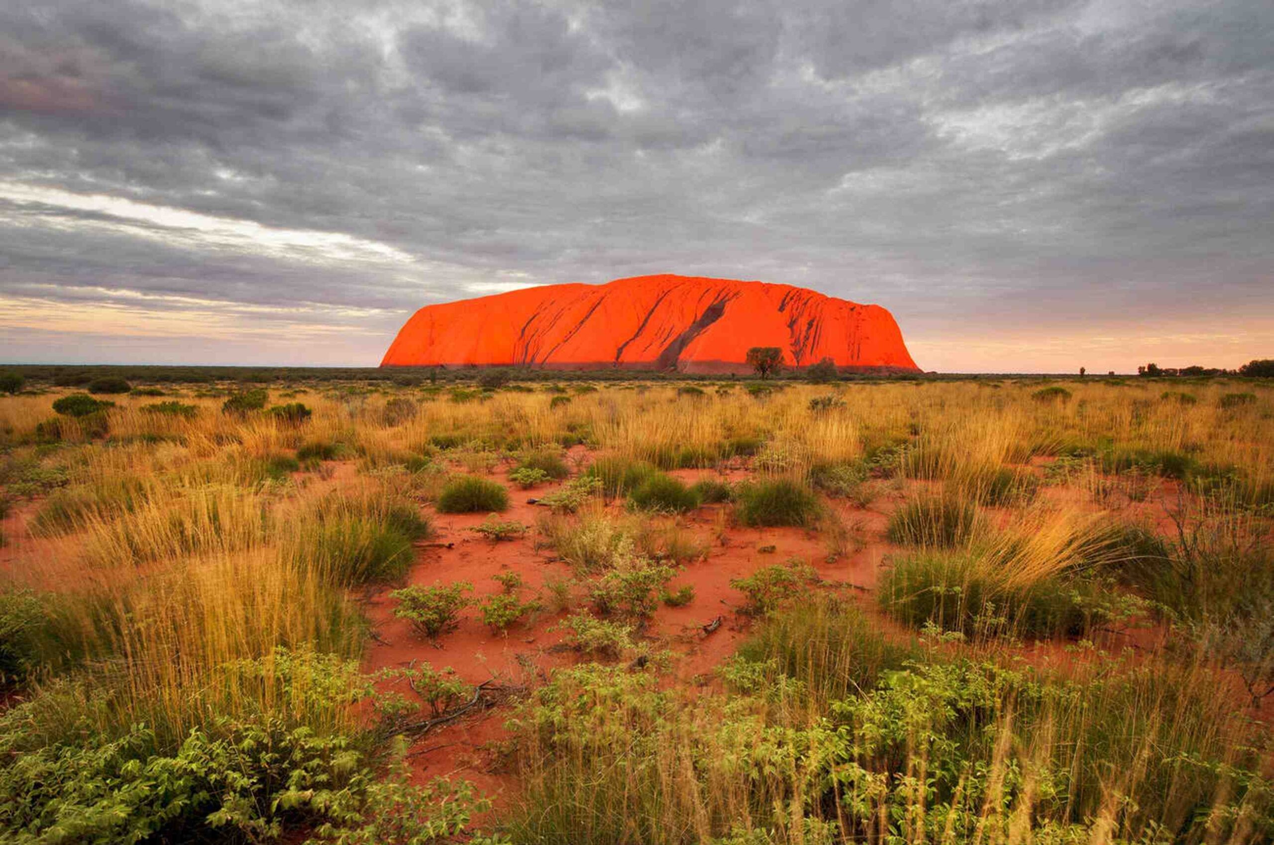 The iconic Uluru rock formation in Australia\u2019s Red Centre, surrounded by desert vegetation under a dramatic sky.